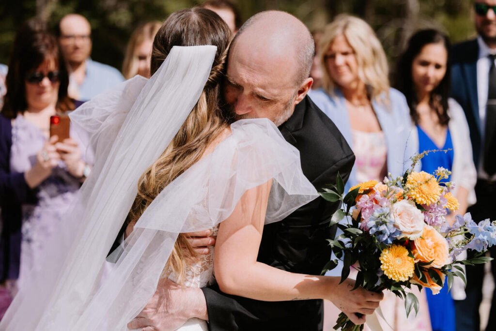 Emotional wedding ceremony moment as the bride embraces her father while holding her bouquet at Silvertip Resort in Canmore, Alberta